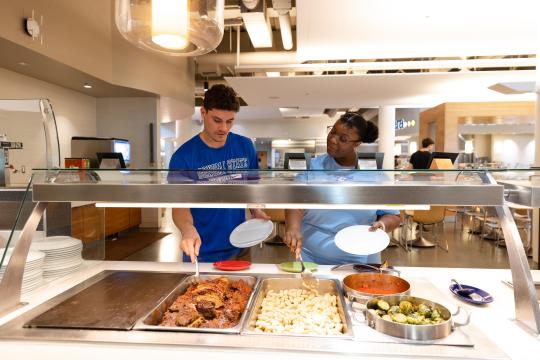 Two students put food onto their plates in the Kitchens