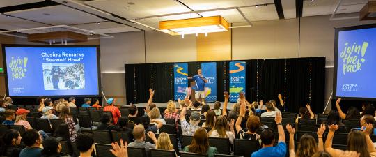 Large audience and a stage with one person and video screens